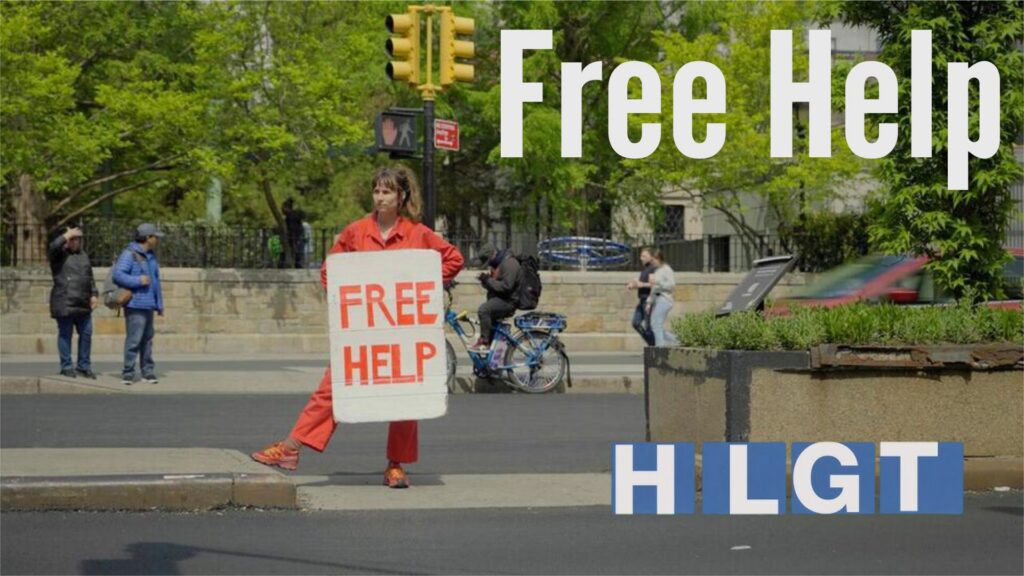 A person in an orange jumpsuit stands at a street corner holding a sign that reads "Free Help". The text "Free Help" is also superimposed on the image in large white letters.